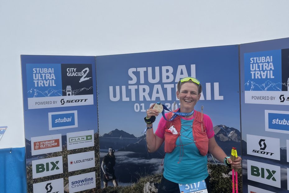 Bianca im Ziel des K32 Stubai Trails mit Finishermedaille vor Sponsorenwand