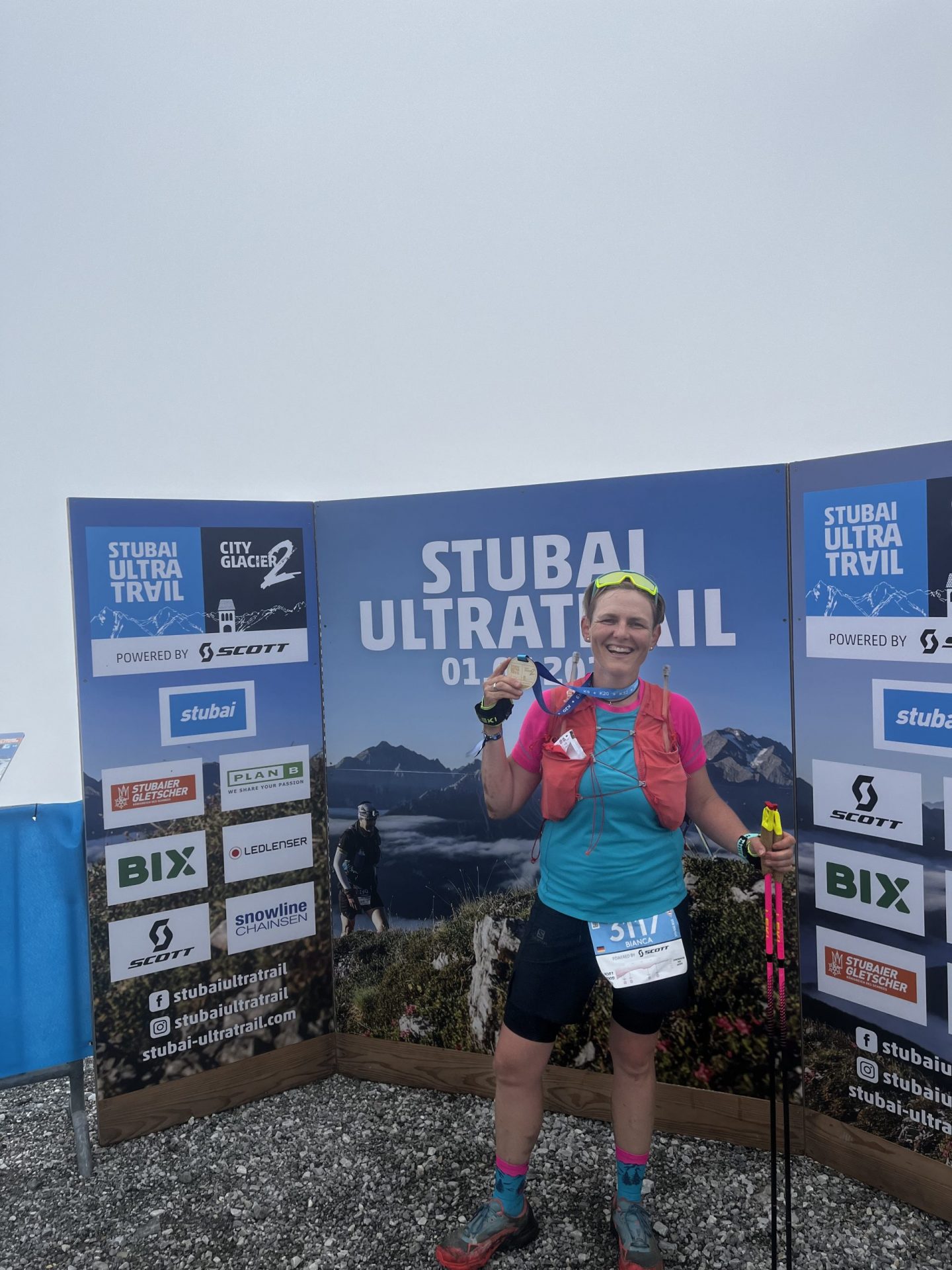 Bianca im Ziel des K32 Stubai Trails mit Finishermedaille vor Sponsorenwand